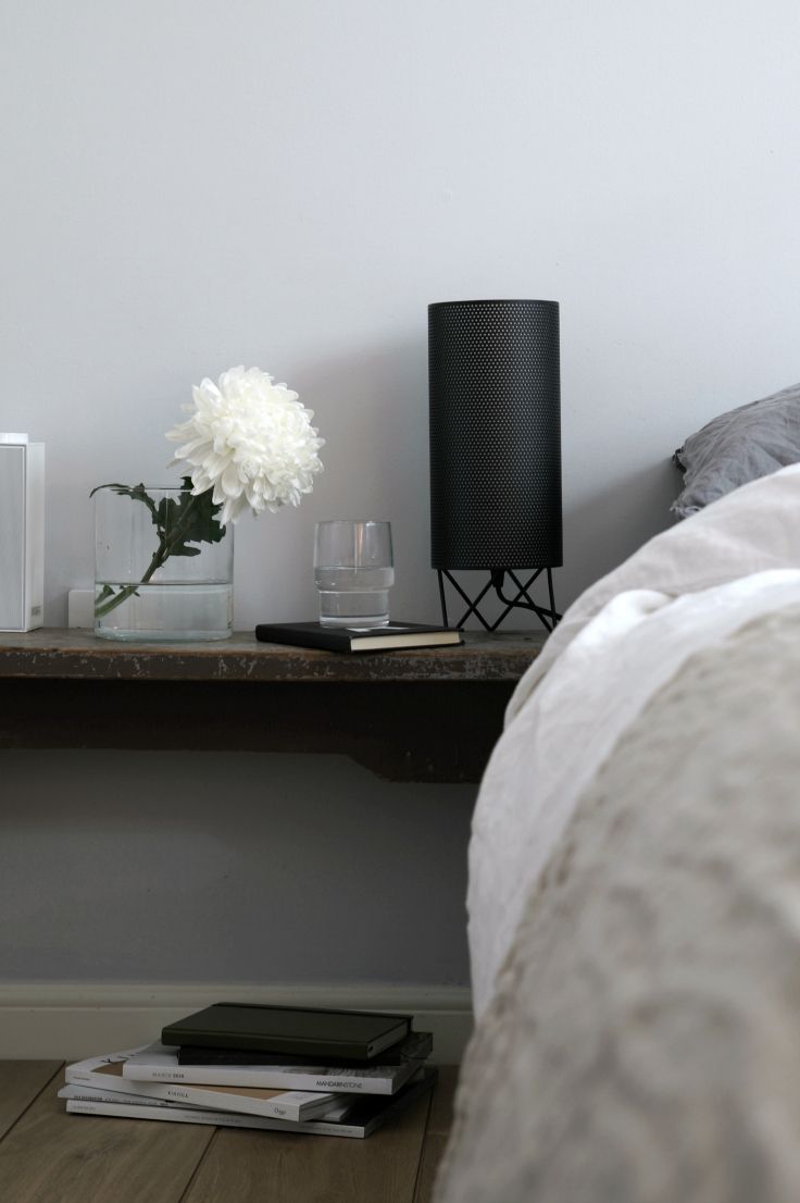 Black cylindrical bedside lamp beside white flowers and clear glasses on a dark shelf next to pale bedding.