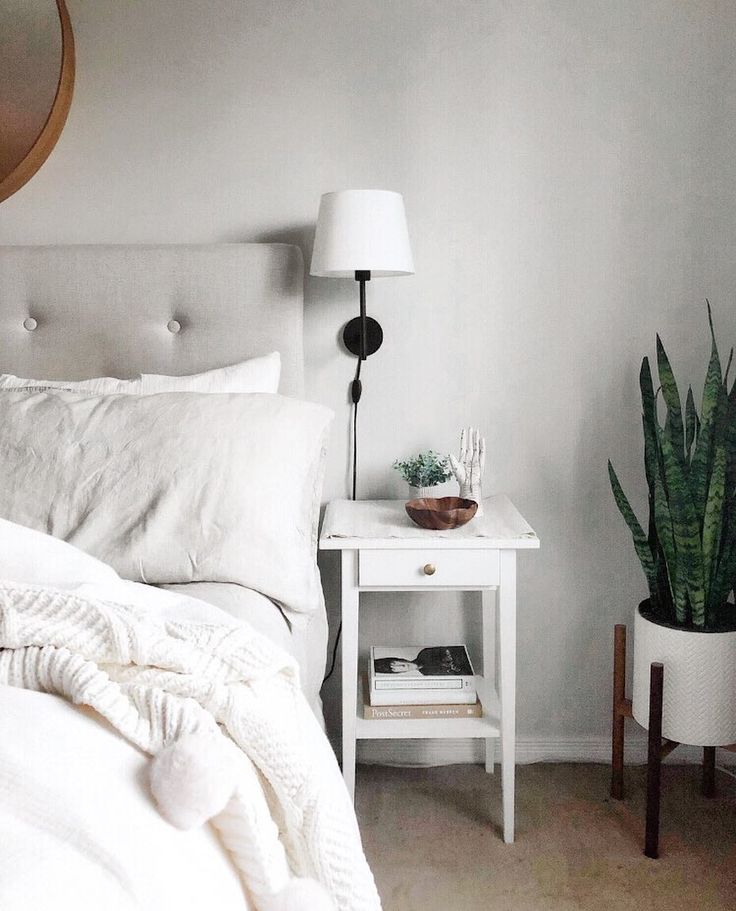 White bedding against a pale gray upholstered headboard beside a white nightstand, black wall light, and tall green plant.