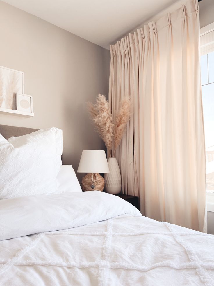 Pale blush curtains beside white bedding, a taupe wall, and a small bedside lamp.