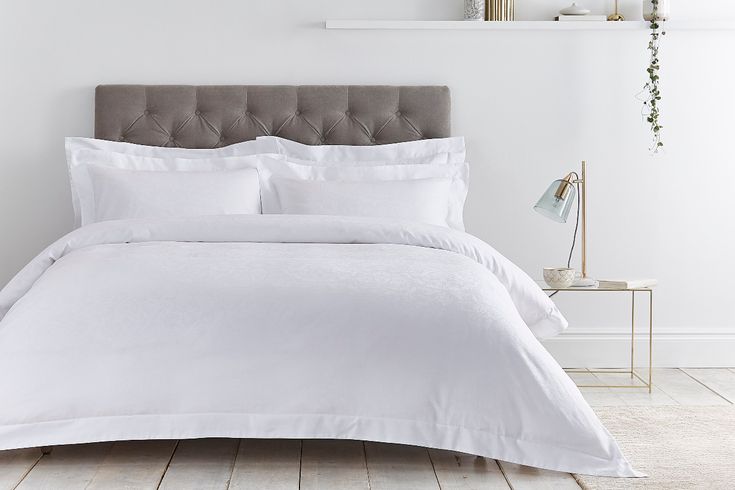 Plain white bedding with a taupe tufted headboard and a slim gold-toned bedside table and lamp.