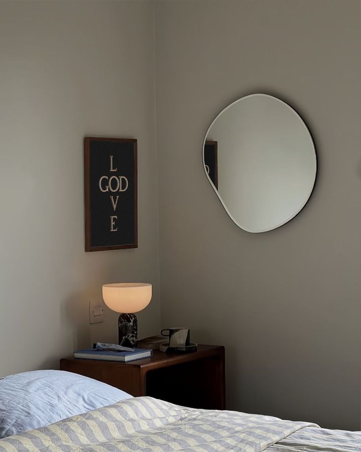 A glowing lamp, books, and a cup on a bedside table below a round mirror.