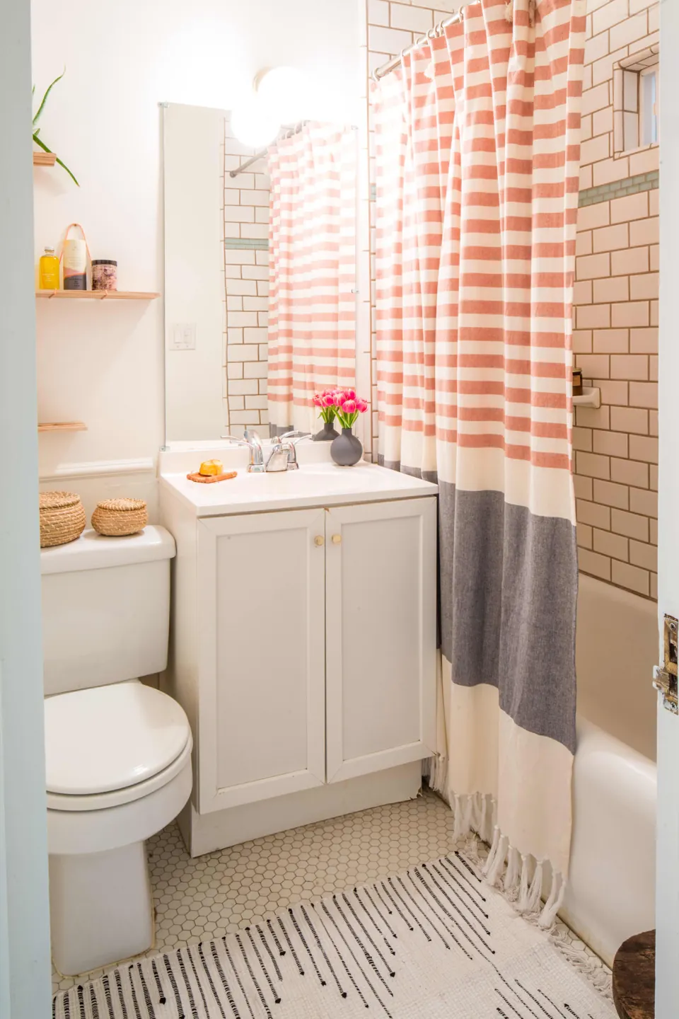 A cream rug with dark lines lies in front of a white vanity and a coral, white, and gray striped shower curtain.