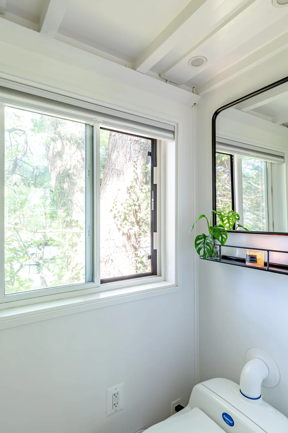 A glass vessel with green leaves and a small candle sit on a narrow mirror shelf beside a bathroom window.