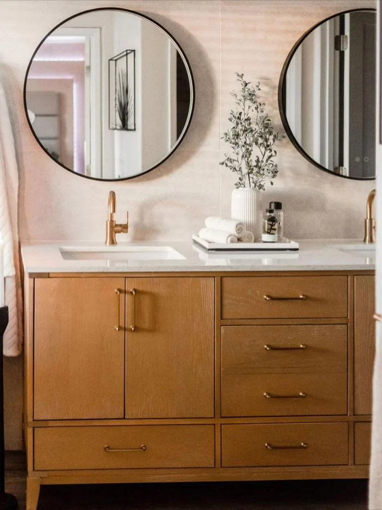 Two black-rimmed round mirrors hang above a wood-toned double vanity with gold-toned faucets and a pale counter.