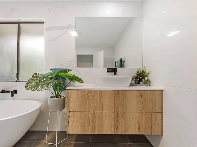 A frameless rectangular mirror hangs above a wood-toned bathroom vanity with a white counter and a dark wall-mounted faucet.