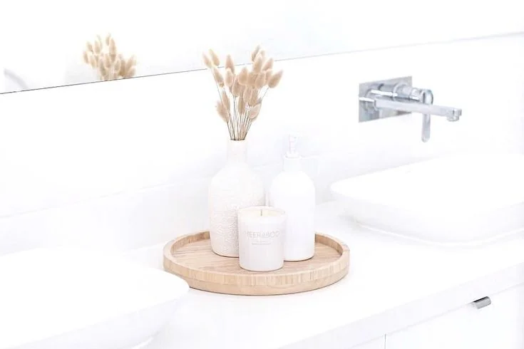 A round wood-toned tray holds a white vase, soap dispenser, and candle on a white bathroom counter.