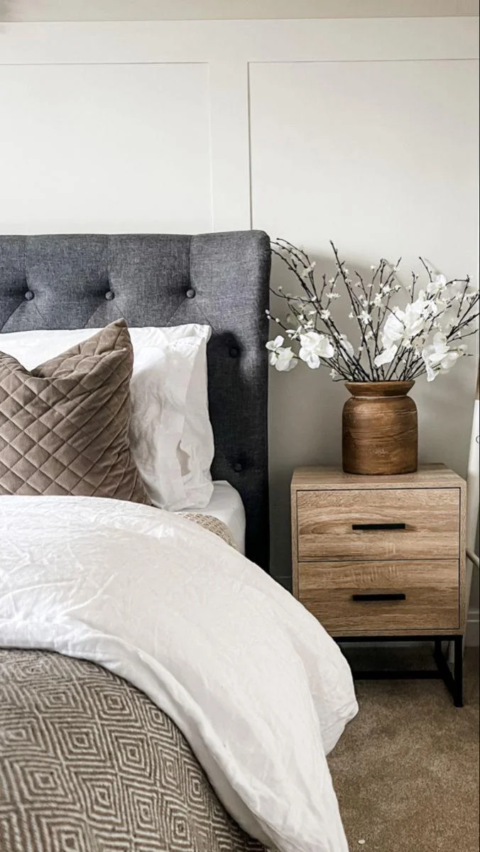 A charcoal headboard frames white bedding beside a brown cushion and a wood-toned bedside cabinet.