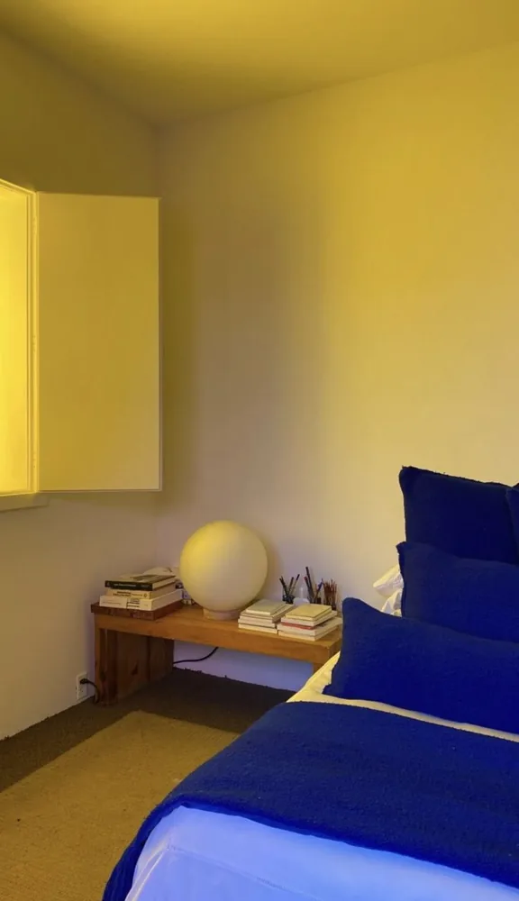 Yellow bedroom walls and ceiling sit above vivid blue pillows and bedding beside a low bedside shelf.