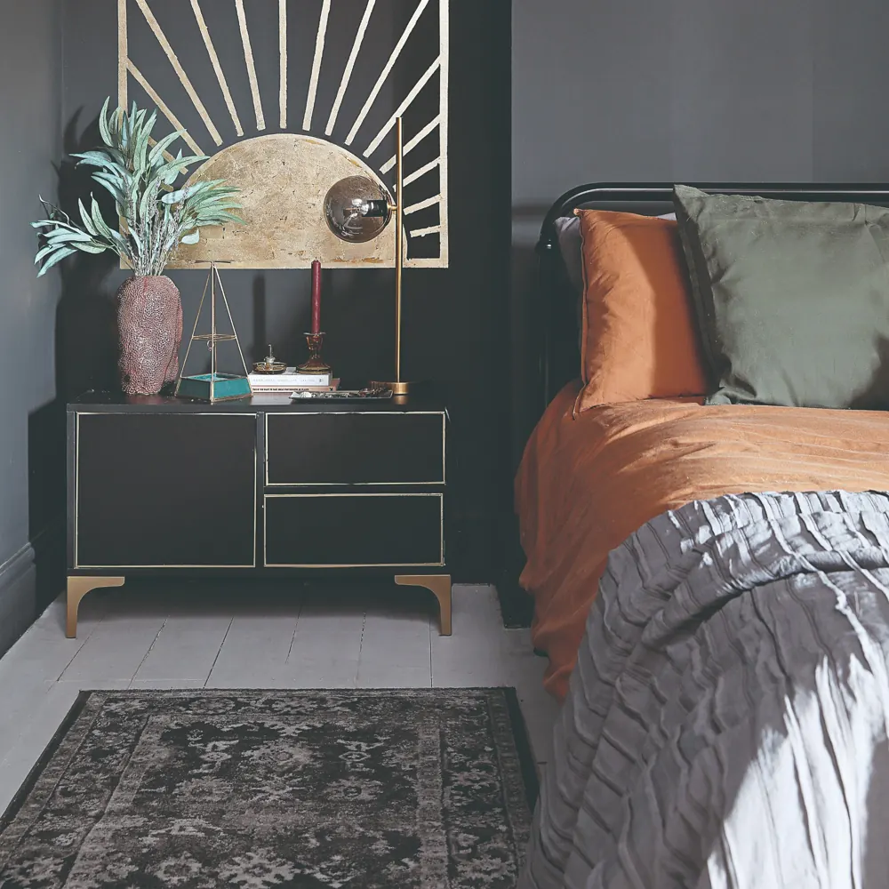 A narrow patterned runner lies beside a bed with rust bedding in a dark bedroom.