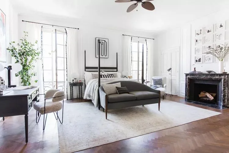 A large pale rug anchors a bed and dark bench in a bright bedroom.