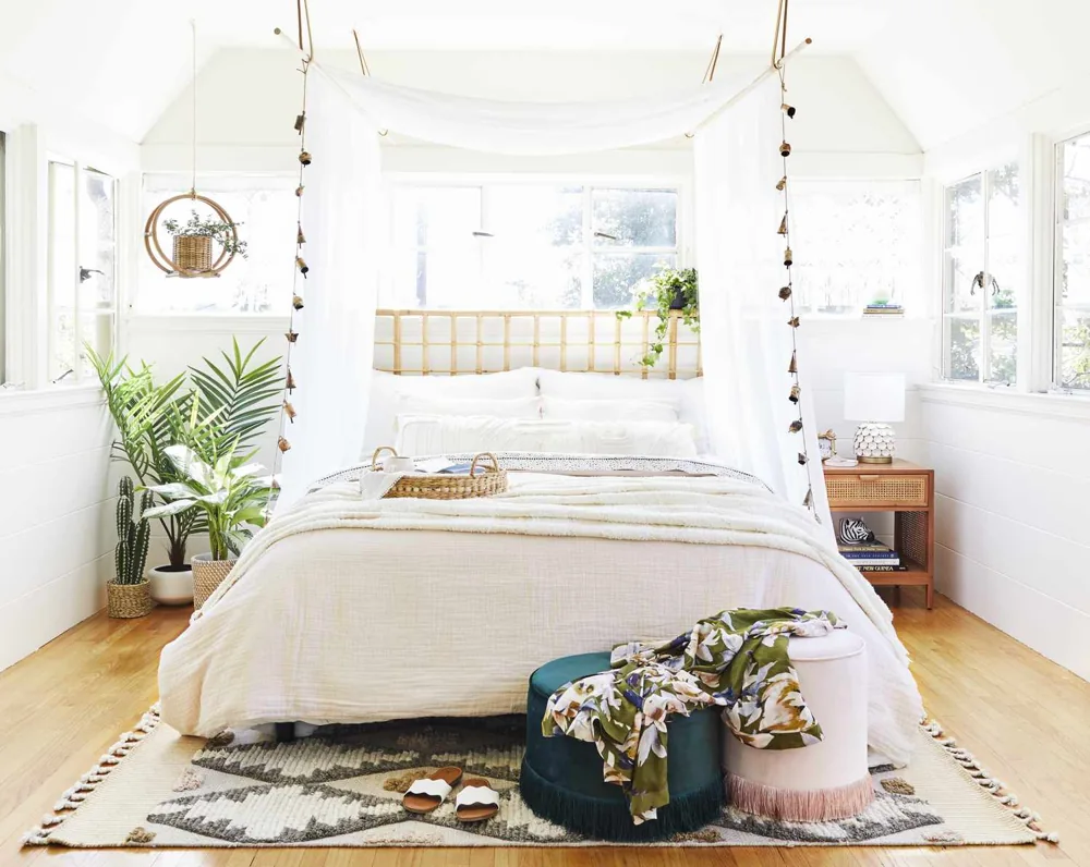 A patterned rug sits beneath a white bed in a bright room with hanging plants.
