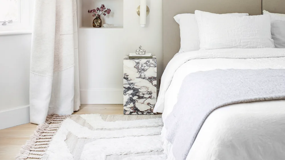 A pale textured rug extends beside a white bed and a dark-veined bedside block.