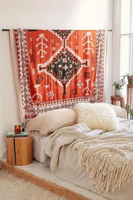 A red patterned wall hanging sits behind a low bed with pale bedding and a fringed throw.