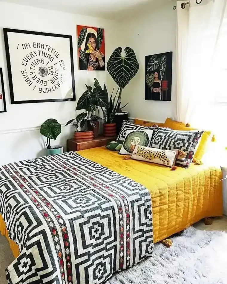 A yellow-covered bed has black-and-white patterned textiles beside potted plants and framed art.