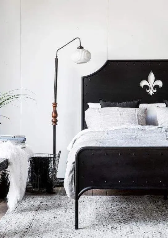 A black metal bed stands beside a floor lamp, pale bedding, a soft chair and a light patterned rug.