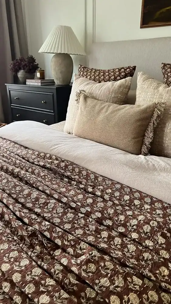 A brown floral bedcover is paired with beige pillows, a pale upholstered headboard and a shaded bedside lamp.