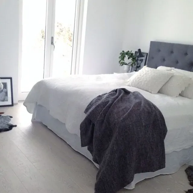 A dark throw lies across white bedding beside a gray upholstered headboard and a bright window.