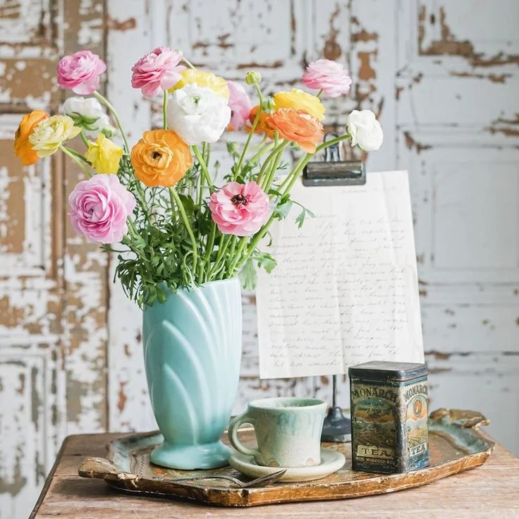 A pale blue vase of colorful flowers stands on a tray with a teacup and a decorated tin.