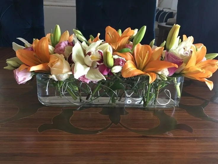A low row of colorful flowers in a long clear container sits on a dining table.