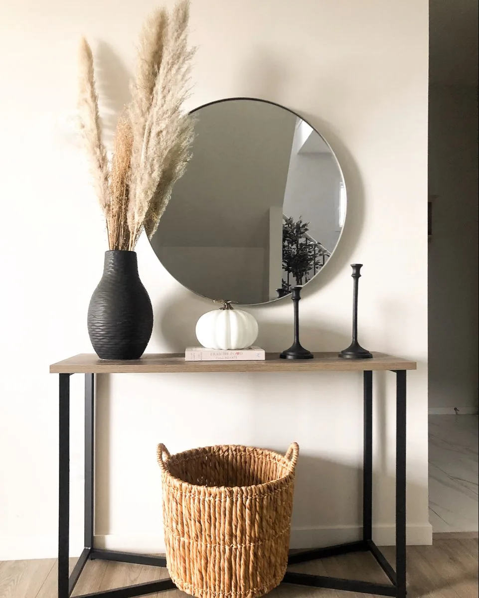 A black vase of pale grasses and a white pumpkin sit below a round mirror on a slim console table.