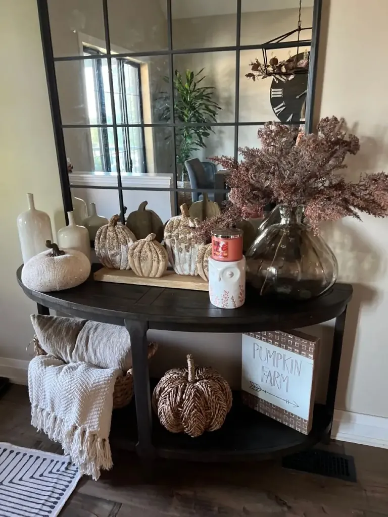 Textured pumpkins, a glass vase of brown stems, and a folded throw decorate a dark curved console table.