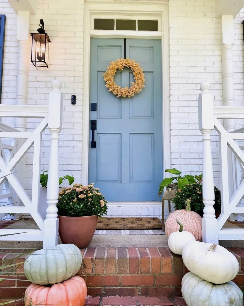Blue front door with a golden wreath, pale pumpkins beside brick steps, and flower pots near the entrance