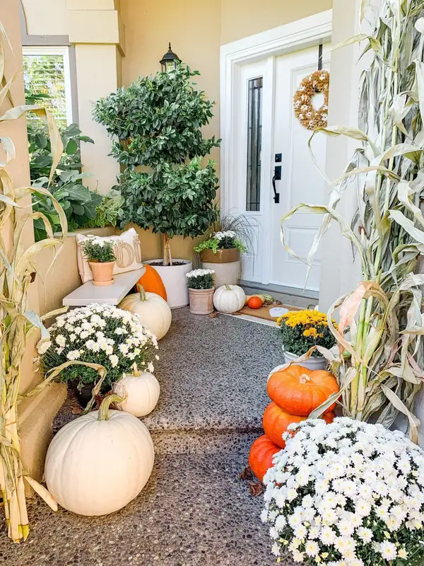 White flowers, pale and orange pumpkins, green foliage, and corn stalks surrounding a porch entrance