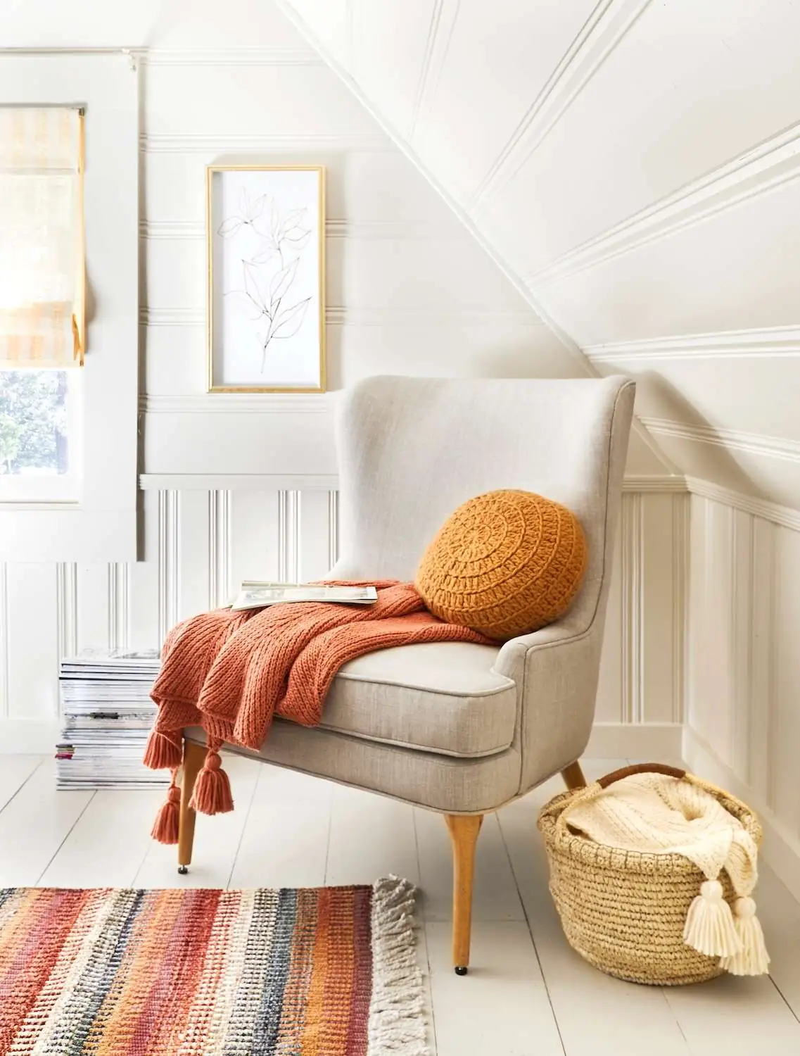 A rust-colored throw and round mustard cushion sit on a pale armchair beside a basket with a cream blanket.