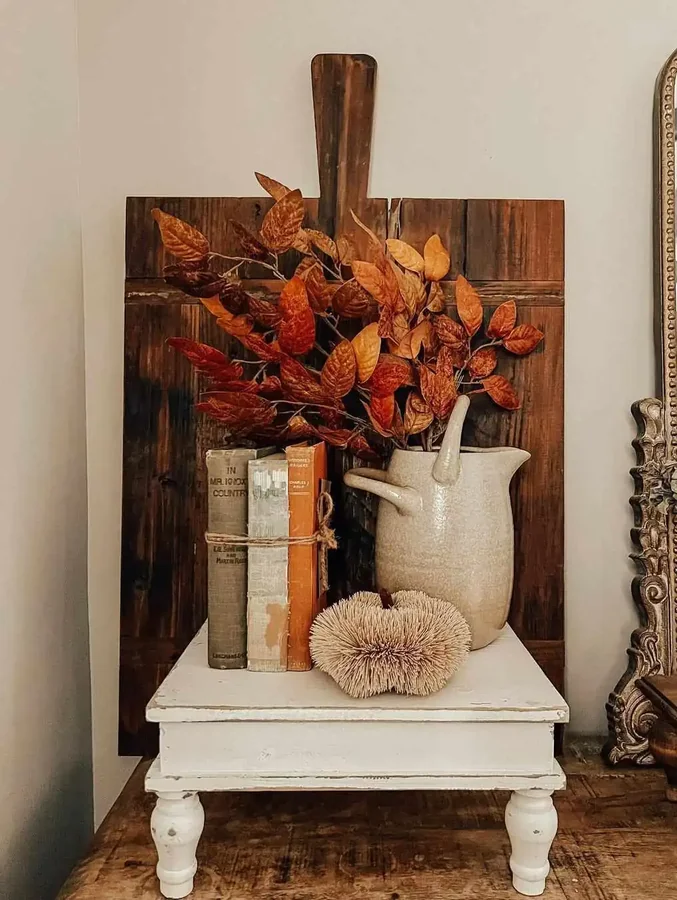 Orange and brown leaves fill a pitcher beside tied books and a small spiky ornament on a white stand.