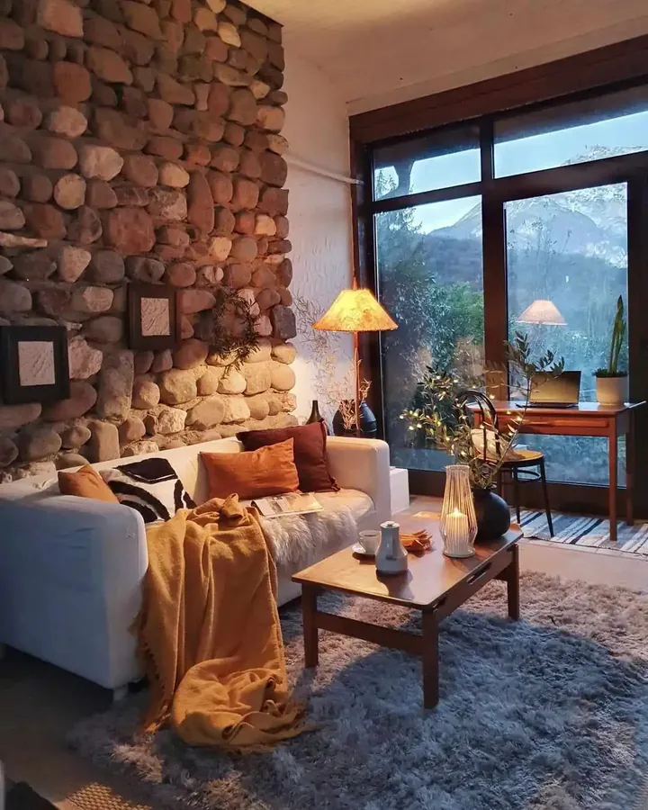 Rust and mustard cushions and a mustard throw brighten a pale sofa beside a stone wall and a wood coffee table.