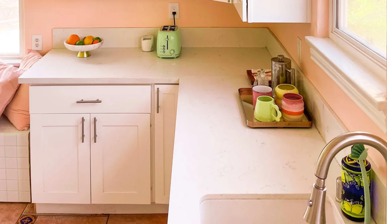 Colorful mugs sit on a tray beside a French press and a kitchen window.