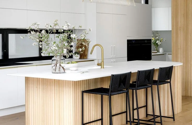 White flowering branches in a clear vase stand toward one end of a pale kitchen island with black stools.