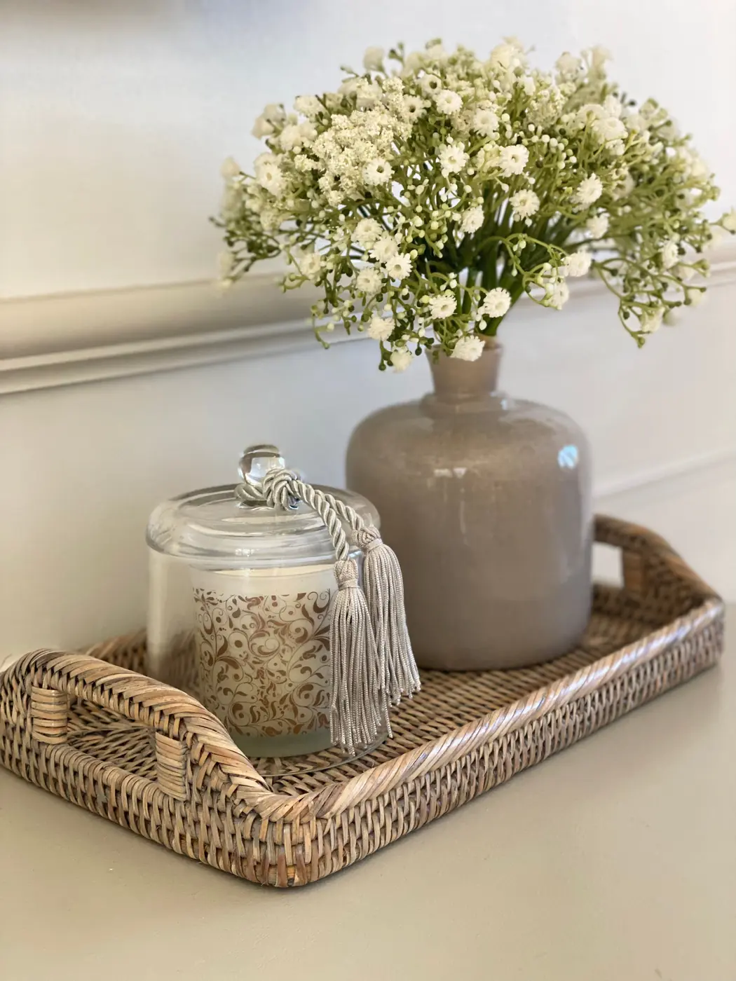 A handled woven tray holds a gray vase of white flowers and a lidded glass jar.