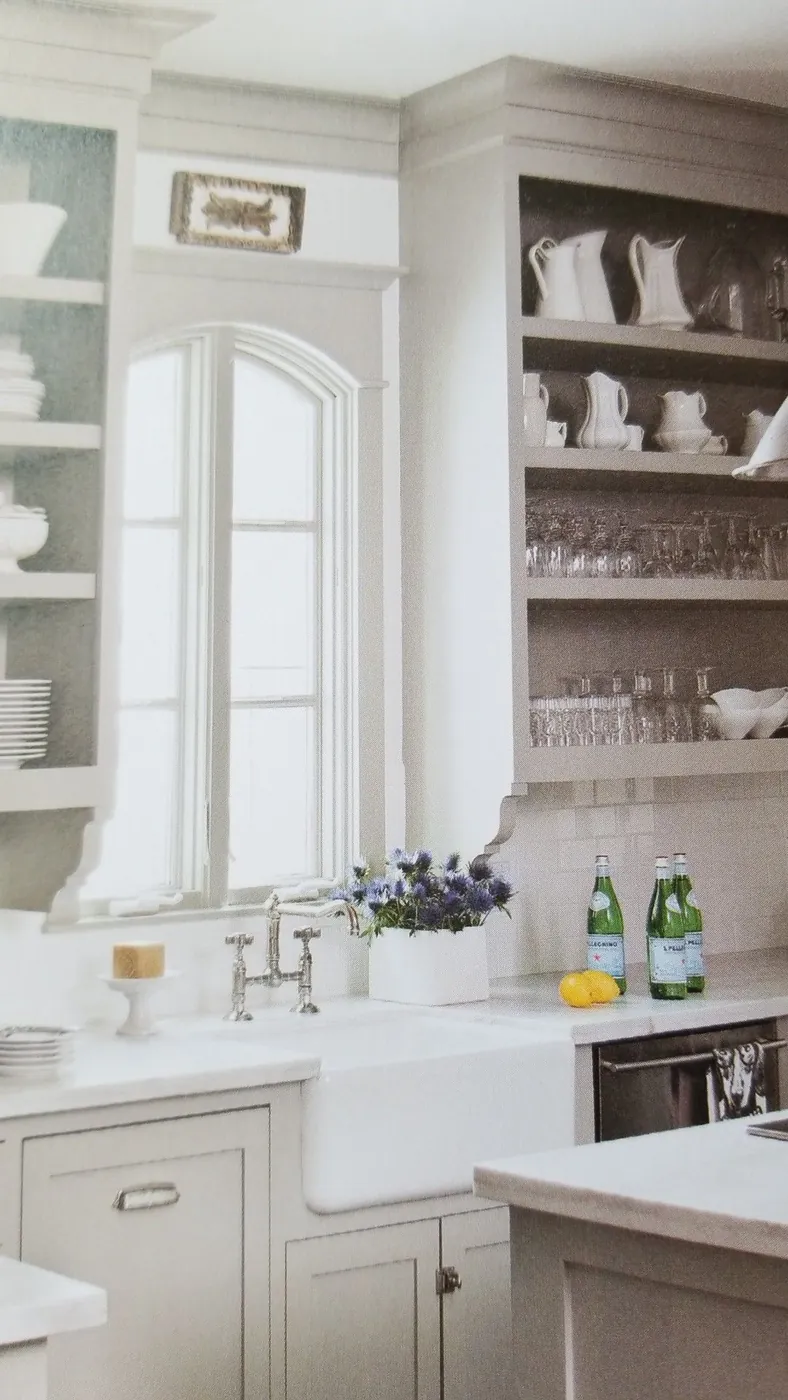 White pitchers and small vessels on kitchen shelves beside an arched window, with rows of glassware below
