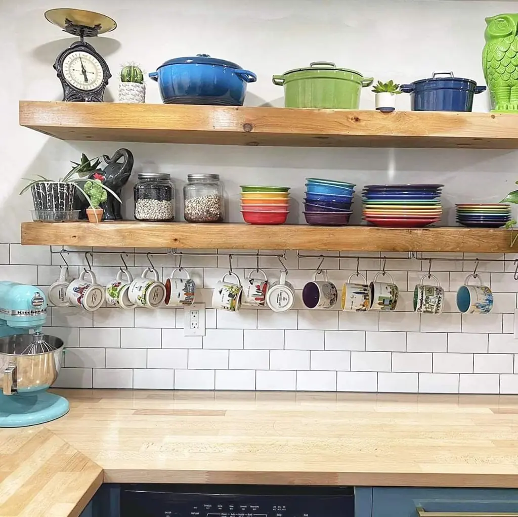 Colorful bowls and plates stacked on wood kitchen shelves beneath blue and green pots, with mugs hanging below