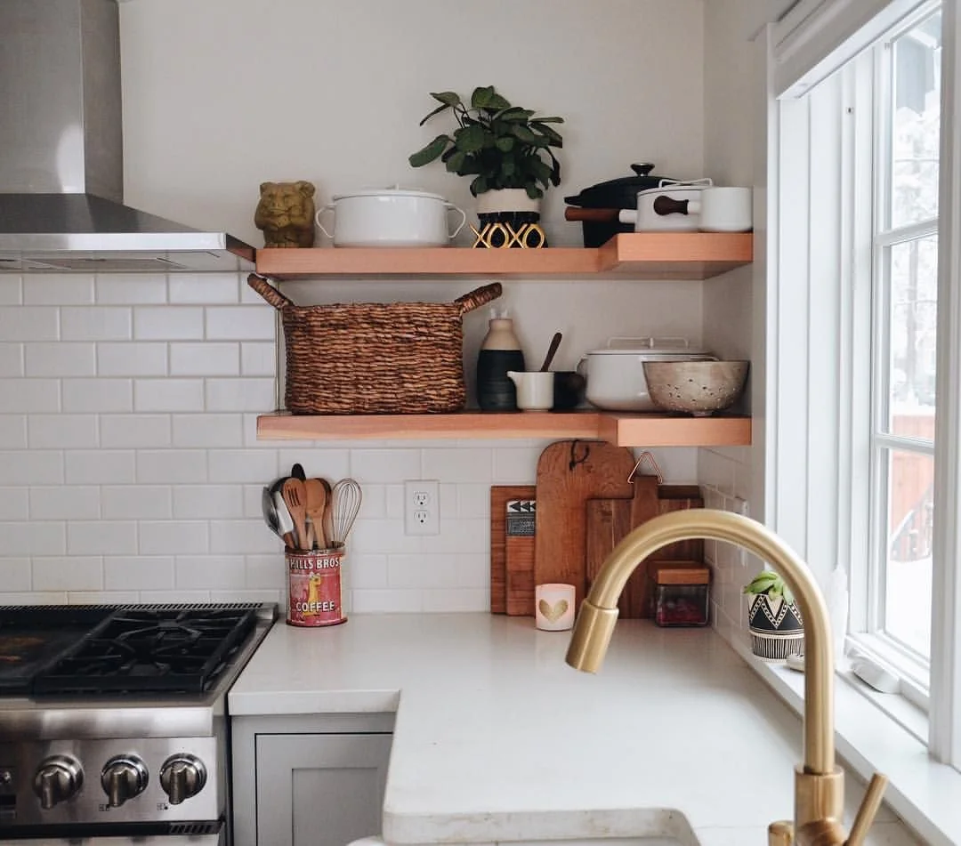 Woven basket beside small vessels and bowls on a lower corner kitchen shelf, with pots and a plant above