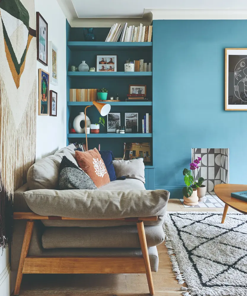 A turquoise wall and shelves sit behind a gray sofa, coral cushion and cream rug with black lines.