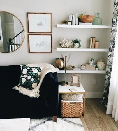 A round mirror, two framed pictures and three white shelves decorate a wall beside a dark sofa.