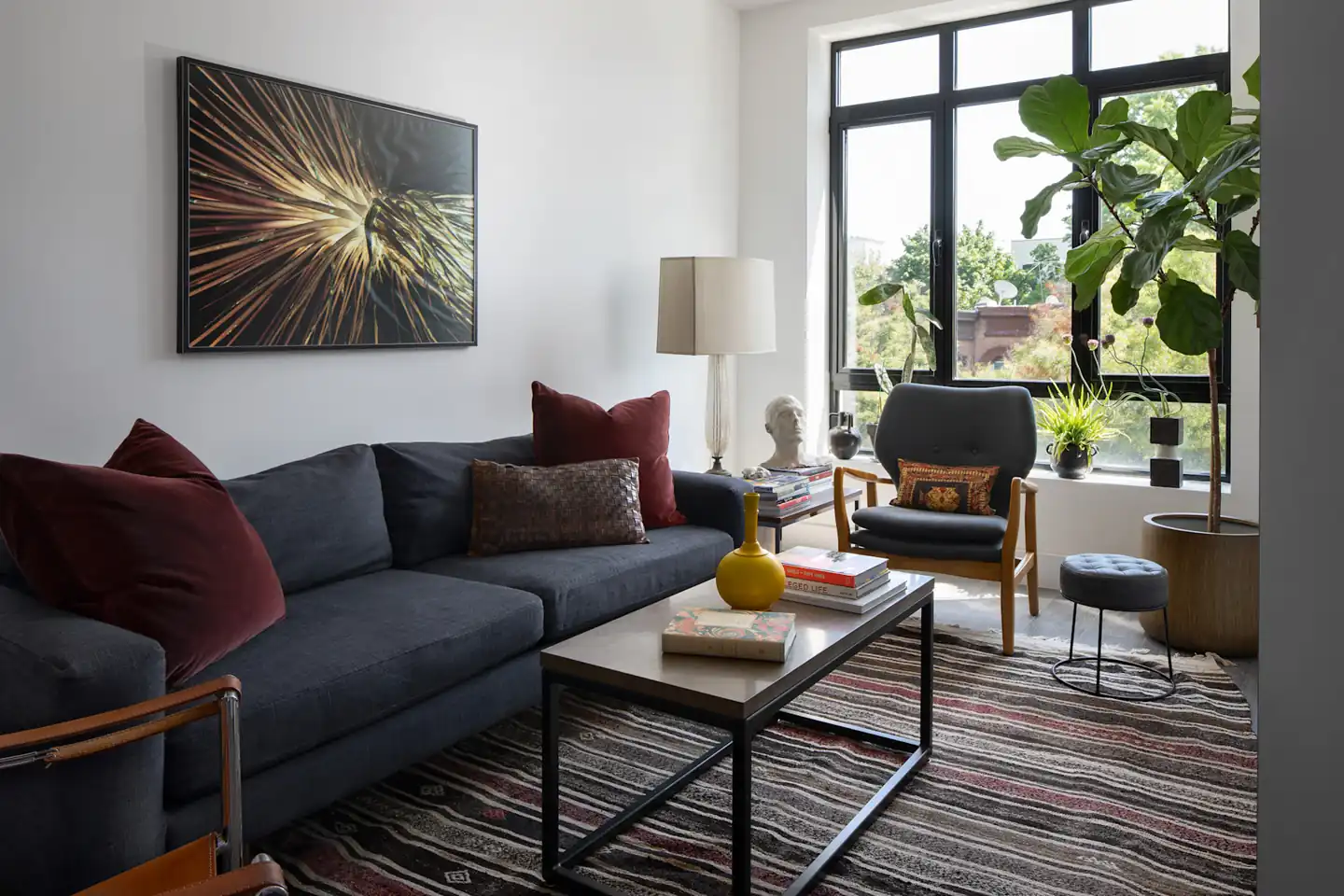 A dark sofa, slim coffee table, and chairs fill a narrow living area by a large window.