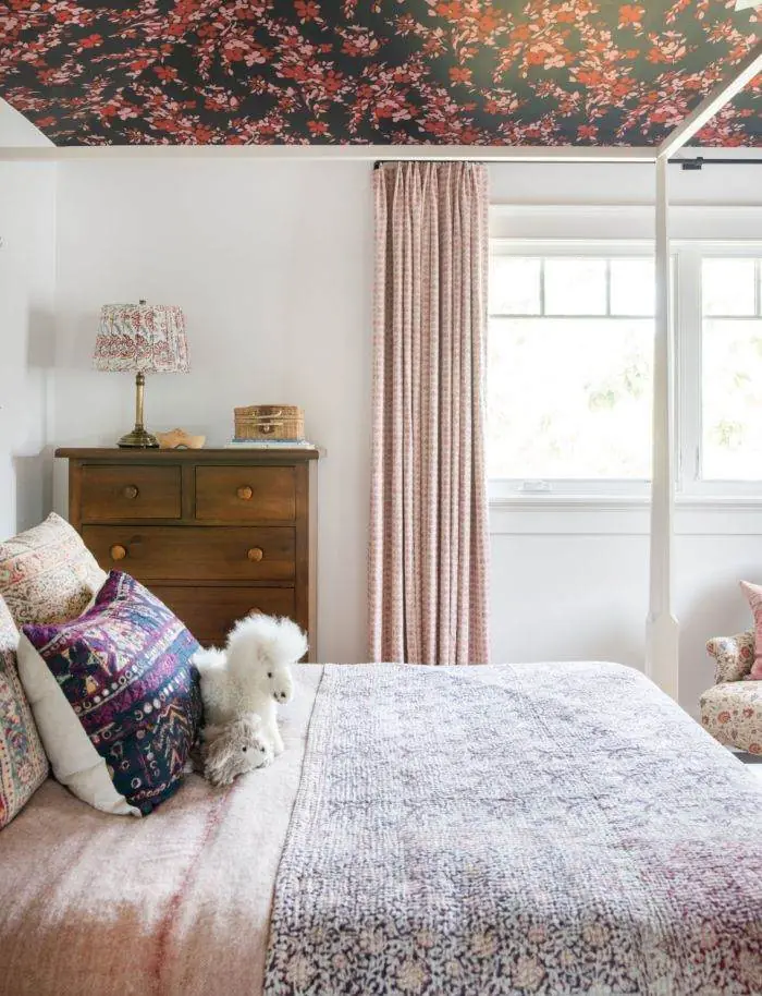 Pink checked curtains beside mixed patterned bedding, white walls, and a dark floral ceiling.