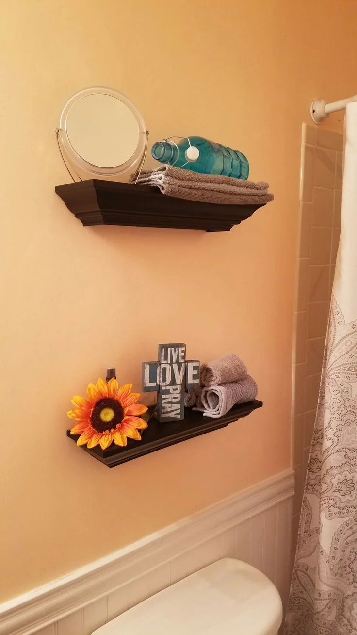 Two dark shelves above a toilet hold rolled towels, a small mirror, a bottle, and a decorative sign.