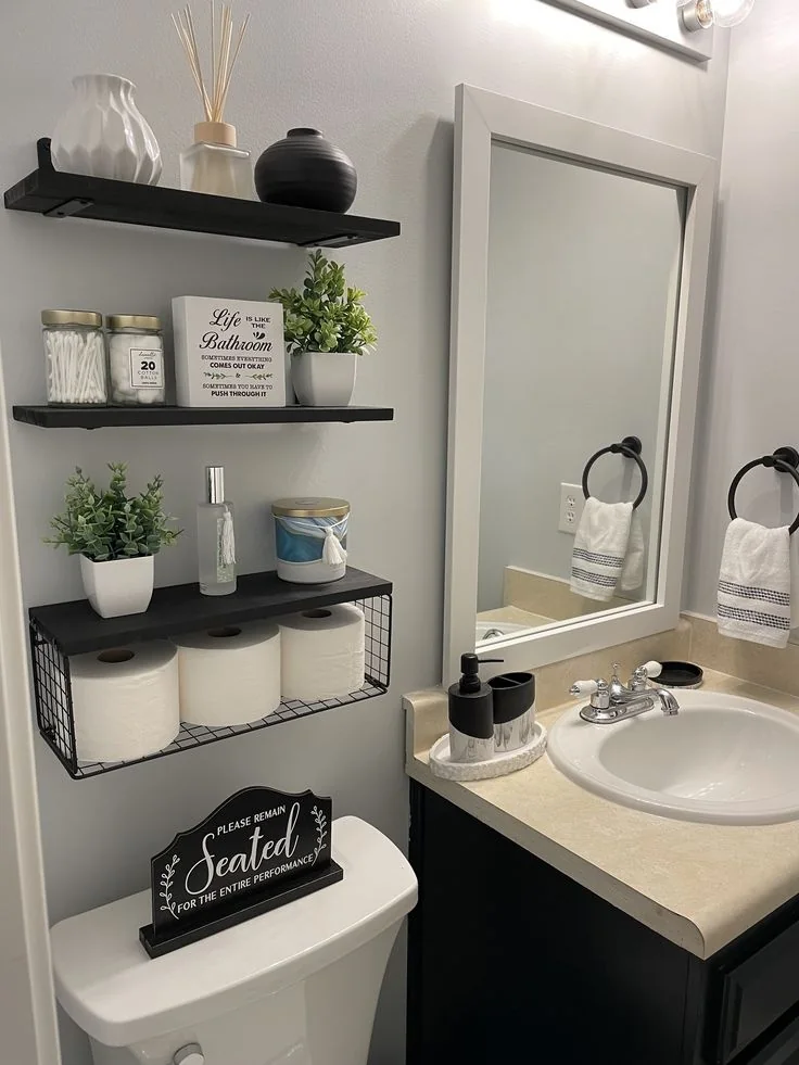 Black wall shelves hold jars, small plants, and bathroom accessories beside a mirror.