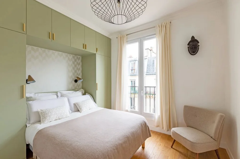 A bed sits in a green cabinet surround beside a tall glazed opening and a chair.