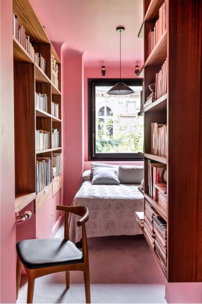 A bed sits beneath a window at the end of a narrow room lined with shelves.