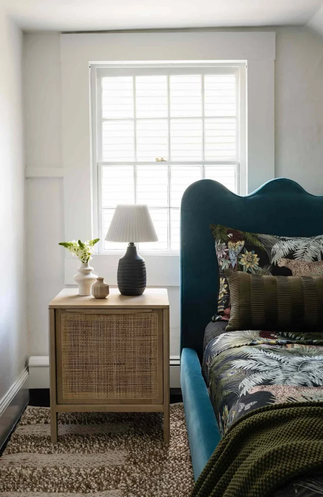 A bed with a curved dark headboard sits off center beneath a window beside a cabinet.