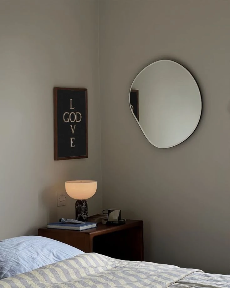 A round wall mirror above a bedside table with a glowing lamp.