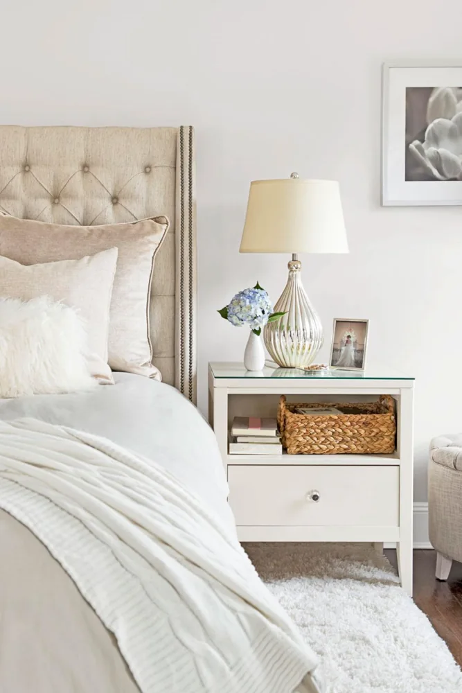 A white bedside cabinet has books and a woven basket on an open shelf above a drawer.
