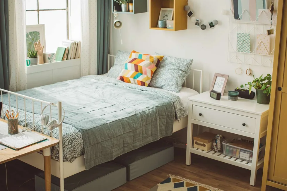 Low gray storage boxes fit beneath a pale bed beside a white bedside unit.