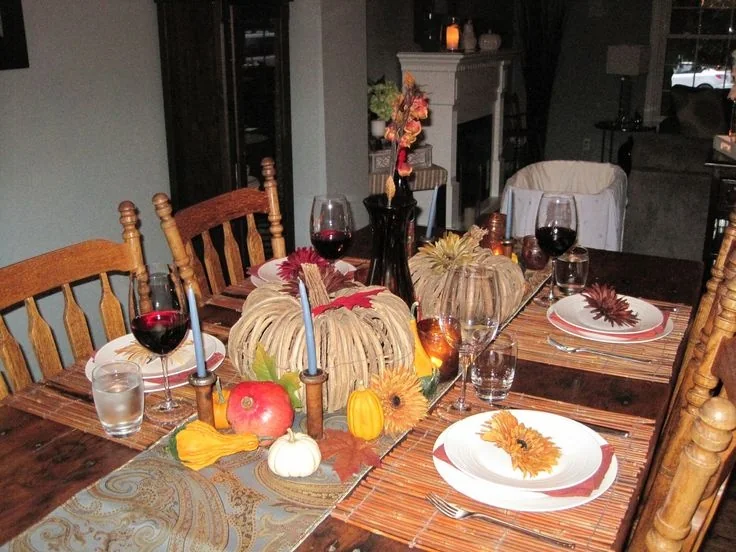 A dining table is set with wine glasses, small pumpkins, fall-colored leaves, and patterned plates.