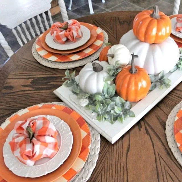 Orange and white pumpkins rest on a white tray beside stacked plates and orange checked napkins.